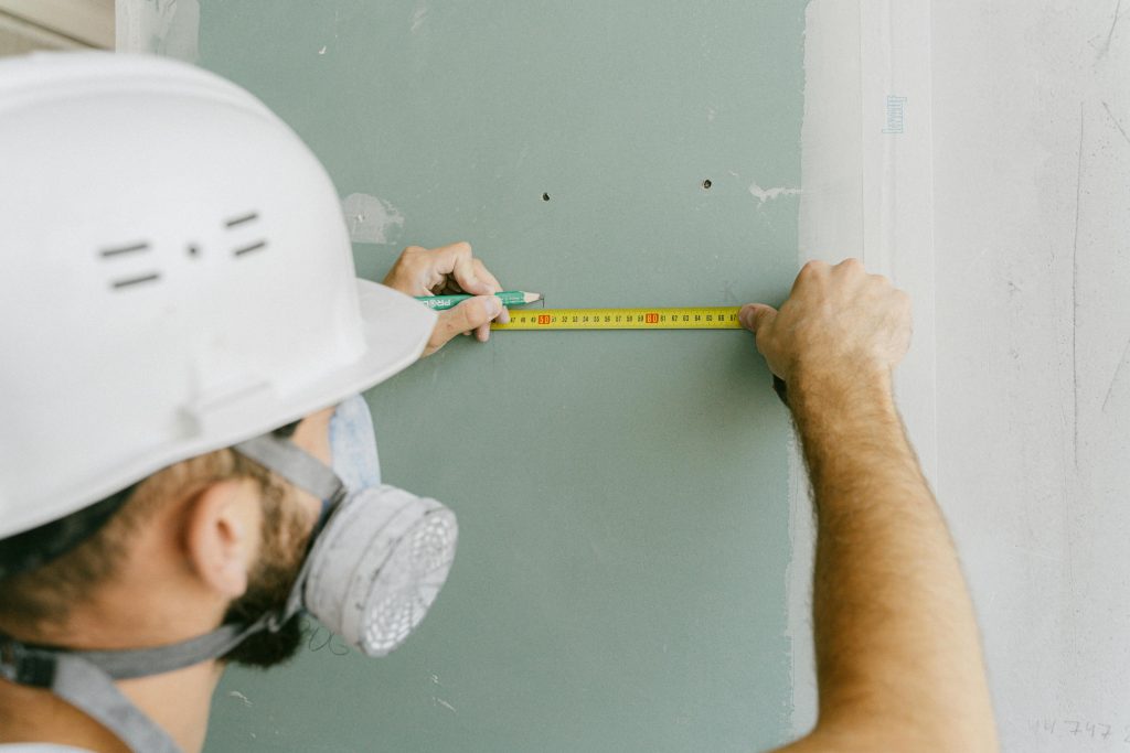 pexels-photo-5493663-5493663 A construction worker precisely measuring drywall, wearing a helmet and mask for safety.
